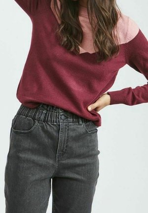 Personne portant un pull à blocs de couleurs rose et bordeaux avec un jean taille haute à taille élastique noire, posant avec une main sur la hanche.
