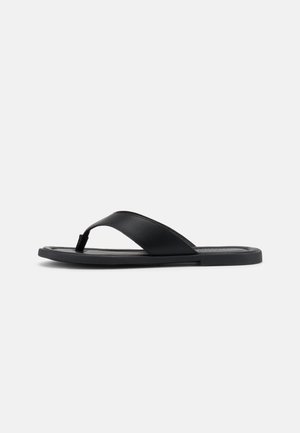 Sandale negre cu talpă joasă, o singură curea largă și șnur între degete, design minimalist, fotografiată pe un fundal simplu gri deschis.