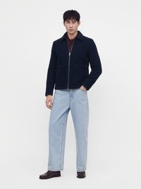 Homme debout portant une veste noire zippée, un jean large bleu clair, des chaussures marron foncé et une chemise rouge foncé sur un fond blanc uni.