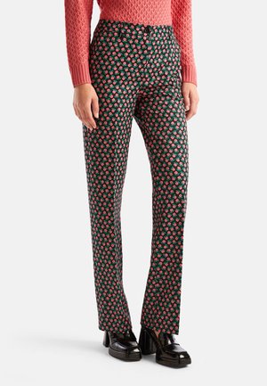 Femme portant un pantalon noir droit avec un imprimé floral rouge, un pull texturé rose et des chaussures à plateforme noires, debout sur un fond blanc.