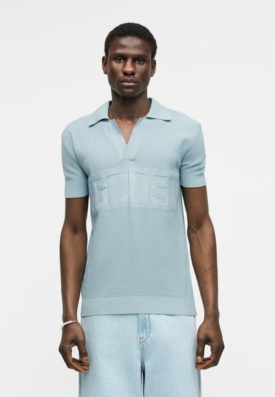 Άνδρας με επάνω φως γαλάζιο πλεκτό πόλο μπλουζάκι με κέντημα λογότυπο "GCDS" και ανοιχτό γαλάζιο τζιν τζιν, στέκεται μπροστά σε λευκό φόντο.