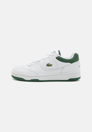 White leather sneakers with a green accent on the heel, featuring a perforated toe box, laces, and the Lacoste logo embroidered on the side.