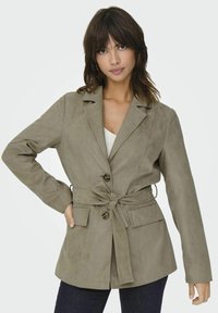 Beige suède blazer met een strikceintuur, twee voorzakken en twee knopen. Heeft een notch-kraag en lange mouwen. Het heeft een stevige textuur.