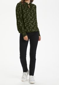 Blouse vert foncé à motifs géométriques, avec un col à nouer et des manches longues, associée à un pantalon noir et des baskets blanches.