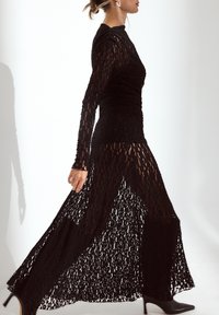 Robe en dentelle noire à manches longues et à corsage ajusté, avec une jupe fluide à fente haute, tissu texturé et détails transparents.