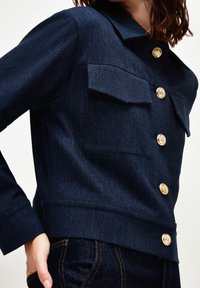 Tummansininen farkkutakki, jossa on kauluksellinen pääntie, kullanväriset napit ja kaksi rintataskua; jämäkkä istuvuus ja lyhyt pituus.