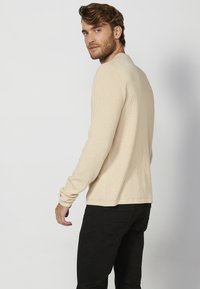 Beige geribbelde gebreide trui met lange mouwen en een ronde halslijn, gedragen met zwarte broeken. De stof heeft een subtiele textuur en een getailleerde snit.