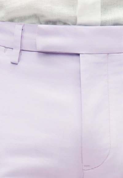 Κοντινό πλάνο στη μέση παντελονιού ανοιχτού μωβ χρώματος με πλαϊνό θηλάκι για ζώνη, επικαλυπτόμενη ζώνη και μπλουζάκι λευκό με υφή που είναι φορεμένο μέσα.