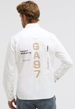 Mann mit hellbraunem Haar trägt weißes Langarmhemd mit beiger Aufschrift "GA 97" und segelthemenbezogenen Texten und Grafiken auf der Rückseite.