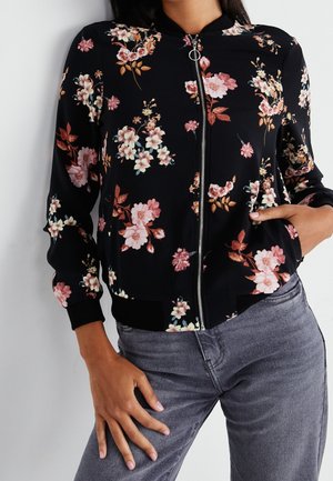 Vrouw draagt een zwarte bomberjack met ritssluiting en een roze-wit bloemmotief, gecombineerd met grijze jeans, met één hand in de zak en de andere op de dij rustend.