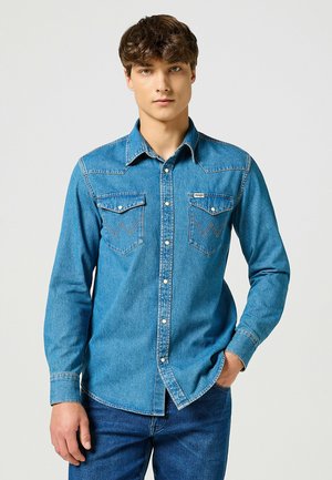 Camicia di jeans azzurro chiaro con maniche lunghe, colletto a punta, due tasche sul petto, bottoni a pressione e cuciture a contrasto. Il indossatore è in piedi.