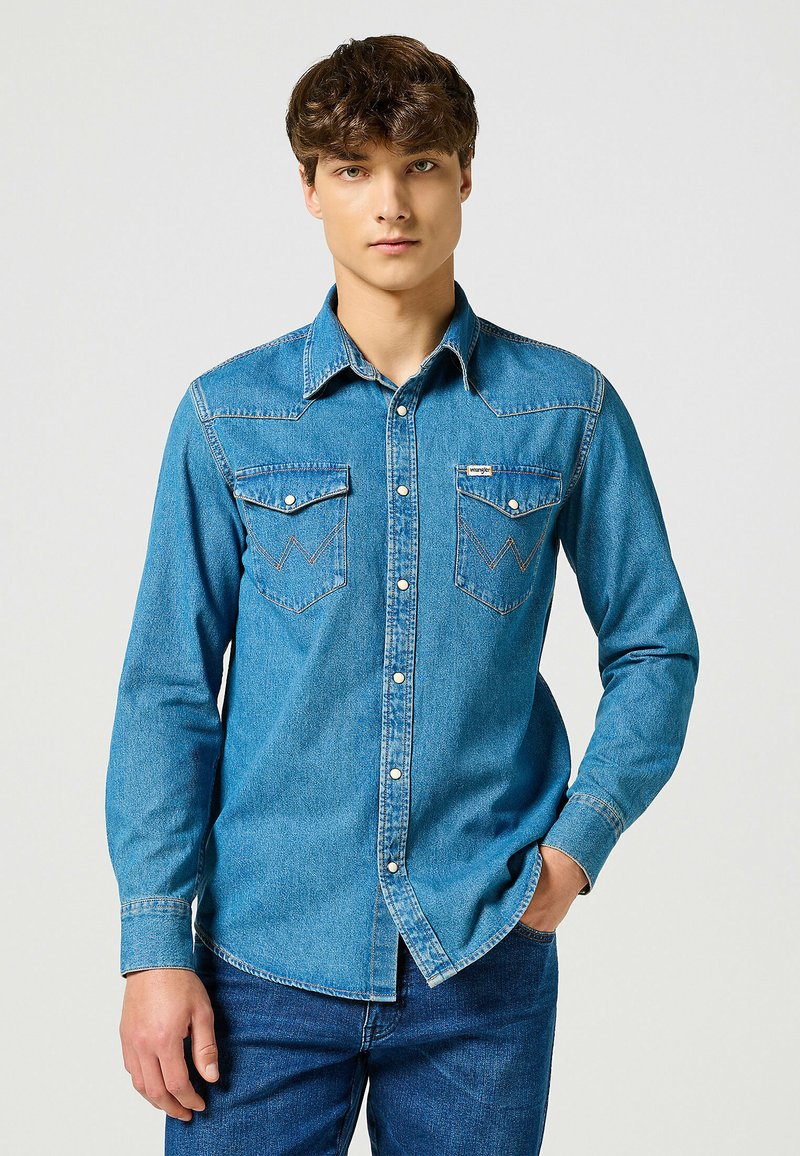 Camicia di jeans azzurro chiaro con maniche lunghe, colletto a punta, due tasche sul petto, bottoni a pressione e cuciture a contrasto. Il indossatore è in piedi.