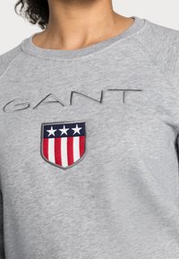 Sudadera gris con un logotipo bordado de "GANT" y un diseño de escudo con rayas rojas y estrellas blancas sobre un fondo azul marino.