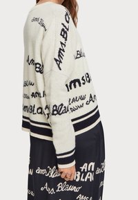 Pull tricoté crème avec motifs de texte noirs ; doté d'un col rond et d'un ourlet rayé. Associé à une jupe sombre arborant un texte similaire.