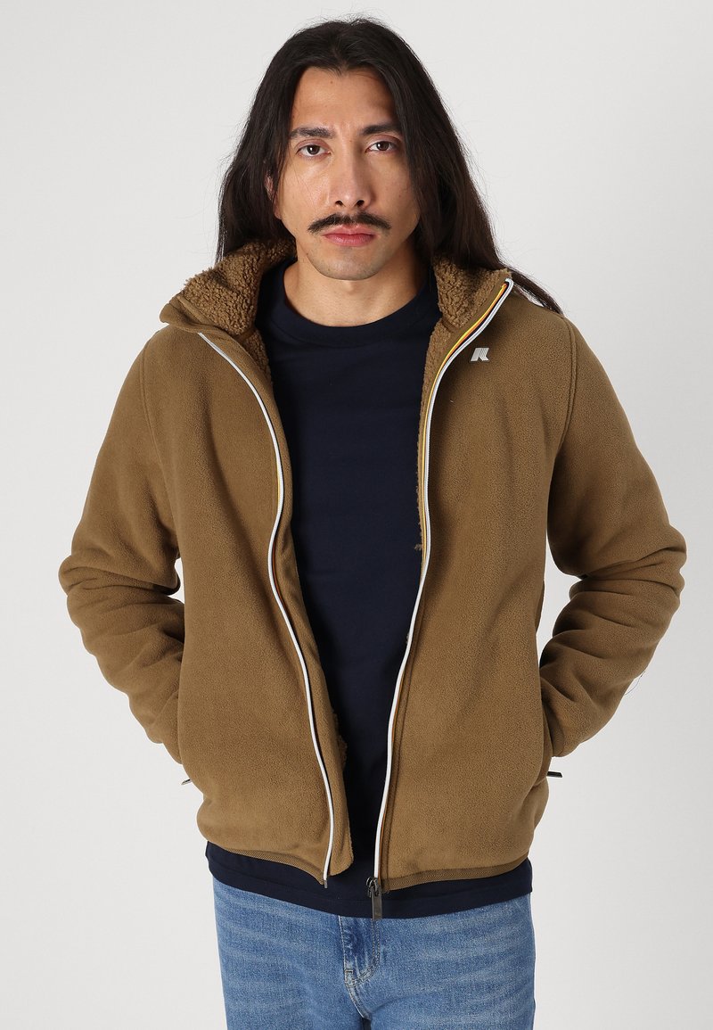 Braune Fleecejacke mit Reißverschluss, strukturiertem Kragen und weißen Akzenten. Getragen über einem navyblauen Hemd, kombiniert mit blauen Jeans.