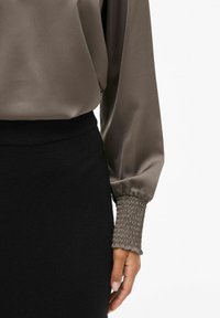 Langærmet bluse i mørk oliven satin med smockede manchetter, parret med en sort tætsiddende nederdel. Glat tekstur og elegant fald.