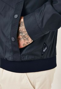 Navy jacket with a smooth, glossy texture, featuring a ribbed navy hem, buttoned placket, and a black triangle logo on the side.