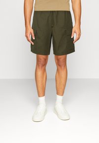 Shorts cargo vert olive en coton, avec poches latérales, taille à cordon et coupe décontractée, assortis à des baskets blanches et des chaussettes blanches.
