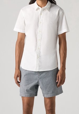 Mand iført en kortærmet hvid skjorte med knapper og blå-hvide lodrette stribede shorts mod en neutral baggrund.