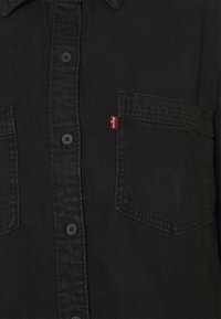 Schwarzes Denimhemd mit spitzem Kragen, sieben Knöpfen, zwei Brusttaschen und einem roten Etikett an der linken Tasche. Strukturierter Stoff.