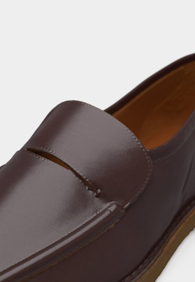 Mocassin en cuir marron avec une bride à boucle, finition lisse et doublure intérieure couleur fauve sur fond blanc.