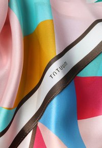 Krāsaina zīda lakatiņa ar drosmīgām ģeometriskām rakstām rozā, zilā, turkīza, dzeltenā un sarkanā krāsā, brūnu apmali un redzamu logotipu.