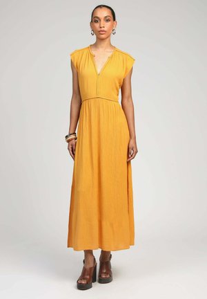 Femme portant une robe longue sans manches jaune moutarde avec un décolleté en V, des fentes latérales, des sandales compensées, des boucles d’oreilles anneaux et des bracelets, debout, de face.