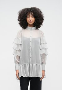 Vaaleanharmaa, läpikuultava pusero, jossa on röyhelöiset hihat, korkea kaulus ja nappikiinnitys edessä. Leveä alareuna, jossa on kerätty helma.