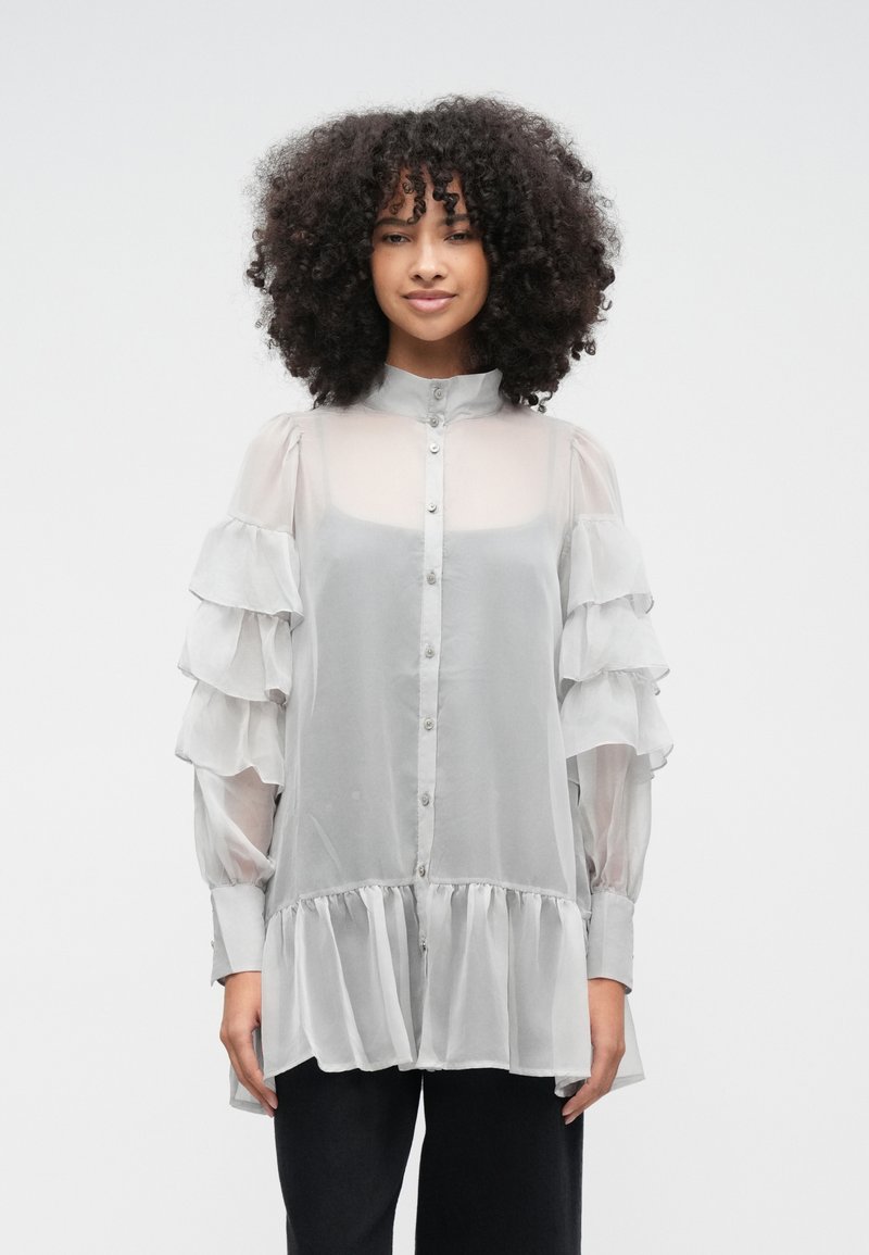 Vaaleanharmaa, läpikuultava pusero, jossa on röyhelöiset hihat, korkea kaulus ja nappikiinnitys edessä. Leveä alareuna, jossa on kerätty helma.