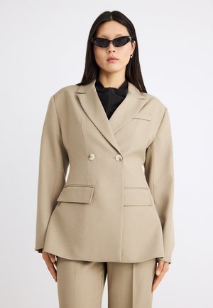 Kvinde iført beige dobbeltradet blazer og matchende bukser med sort top og smalle sorte solbriller mod hvid baggrund.