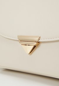 Beigen värinen keinonahkainen laukku, jossa on etuosassa kolmionmuotoinen kultahelatakiinnike, siistit linjat ja hienovaraiset ompelukoristeet.