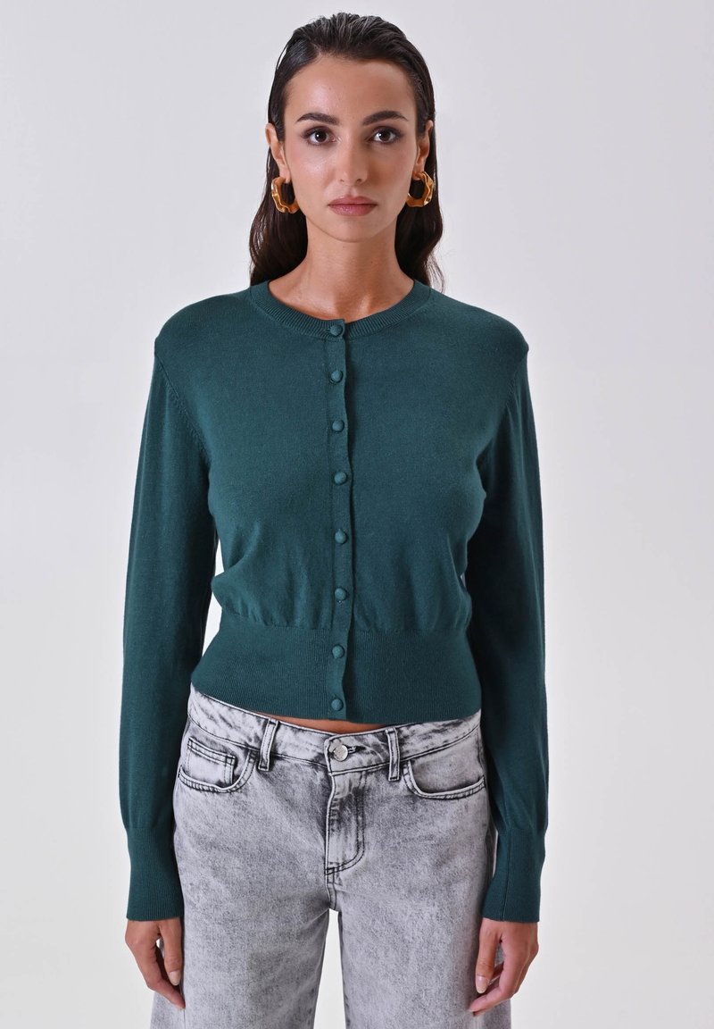 Grüne gestrickte Strickjacke mit langen Ärmeln, Rundhalsausschnitt und Knopfverschluss. Weicher Stoff und taillierte Passform. Getragen mit hellgrauen Jeans.