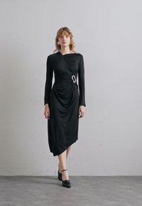 Vestido negro de manga larga con un diseño drapeado, que cuenta con un detalle de hardware plateado en la cintura y un dobladillo ligeramente asimétrico.