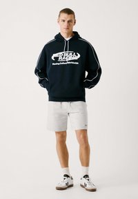 Sudadera con capucha navy con el logo blanco de "Red Bull Racing", combinada con pantalones cortos gris claro y zapatillas blancas con detalles en negro.