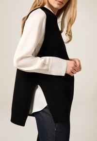 Czarny dziany sweter bez rękawów z bocznymi rozcięciami, noszony na białej koszuli z długim rękawem. Posiada okrągły dekolt i delikatną prążkowaną fakturę.