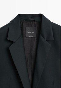 Mörkgrön kavaj med slagkrage, texturerad tyg, en inre etikett och slank design, vilket visar en skräddarsydd look med minimala accenter.