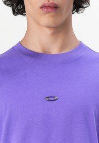 Purple cotton t-shirt featuring a round black metal logo detail on the chest, with a crew neckline and a smooth fabric texture.