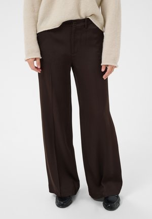 Personne portant un pantalon marron foncé à jambes larges, un pull beige et des chaussures noires, debout devant un fond uni.