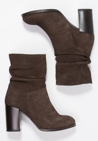 Botines de ante marrón con un diseño arrugado, que presentan un tacón cuadrado y un acento de tacón negro liso. Textura suave sin patrones visibles.