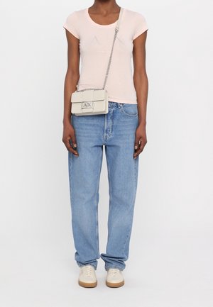 Person trägt ein hellrosa kurzärmeliges Shirt, blaue Jeans, weiße Sneaker und eine kleine beige Umhängetasche mit Kettenriemen und Metall-Logoschließe.