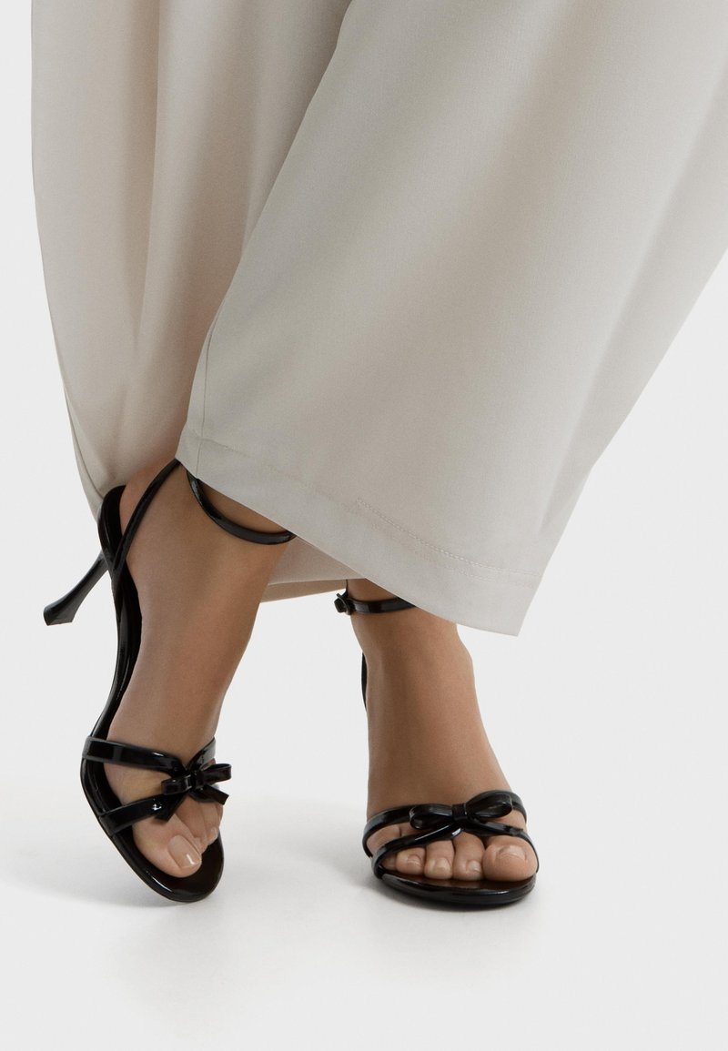 Picioare încălțate cu sandale cu toc înalt din piele lacuită neagră, cu curele la gleznă și funde, asortate cu pantaloni largi bej deschis.