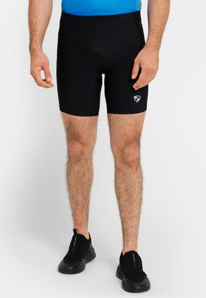 Mann trägt schwarze enganliegende Sportshorts und schwarze Slip-On-Sneaker, steht vor einem schlichten weißen Hintergrund.