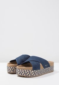 Plattformssandaler med flätade blå och beige sulor och korsade jeanstövlar på en vit yta med en snar bakgrund.