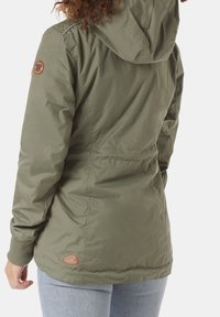 Giacca leggera color verde oliva con cappuccio, polsini elasticizzati e vita regolabile con cordino. Presenta una toppa testurizzata sulla spalla.