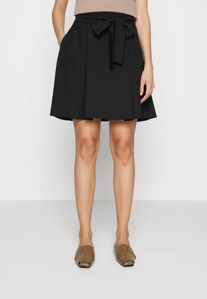 Mujer con falda negra hasta la rodilla con cinturón atado y zapatos planos abiertos de color marrón, de pie contra un fondo liso.