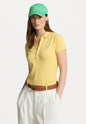 Yellow polo shirt with a turquoise emblem, paired with cream pants and a brown woven belt. A green cap completes the look.