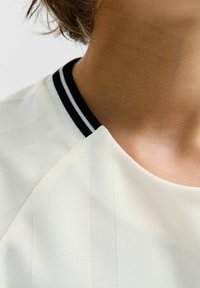 Primer plano del cuello y el hombro que lleva una camisa blanca con un cuello a rayas blanco y negro y cabello corto castaño.