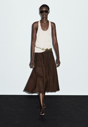 Model wears a beige tank top and a long, chocolate brown skirt. The skirt features a flowy design and is complemented by a gold belt.