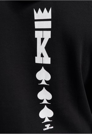 Musta puuvillane T-särk, millel on vertikaalne valge graafika kroonist, tähe "K" ja ruutmängusümbolitest, allpool tekst "KING OF COMPTON".