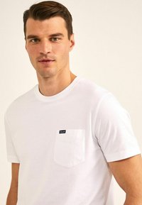 Witte katoen t-shirt met een ronde hals, korte mouwen en een borstzak. De zak heeft een klein merklabel. Soepele textuur.